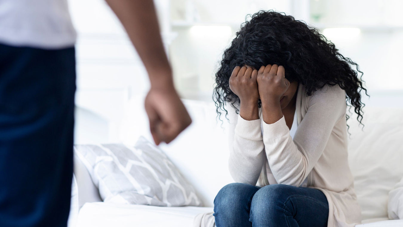 A woman with curly black hair sits on a white couch, covering her face with her hands, while a man in a white shirt and dark pants stands blurred in the foreground.