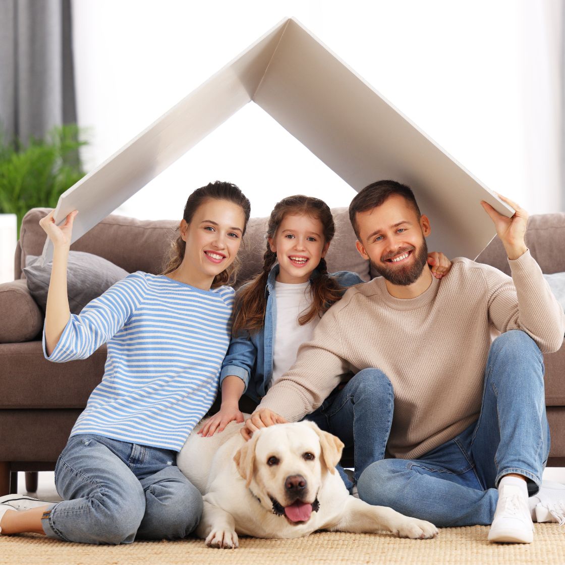 Family of three with a dog sitting on the floor, forming a house shape with their hands.