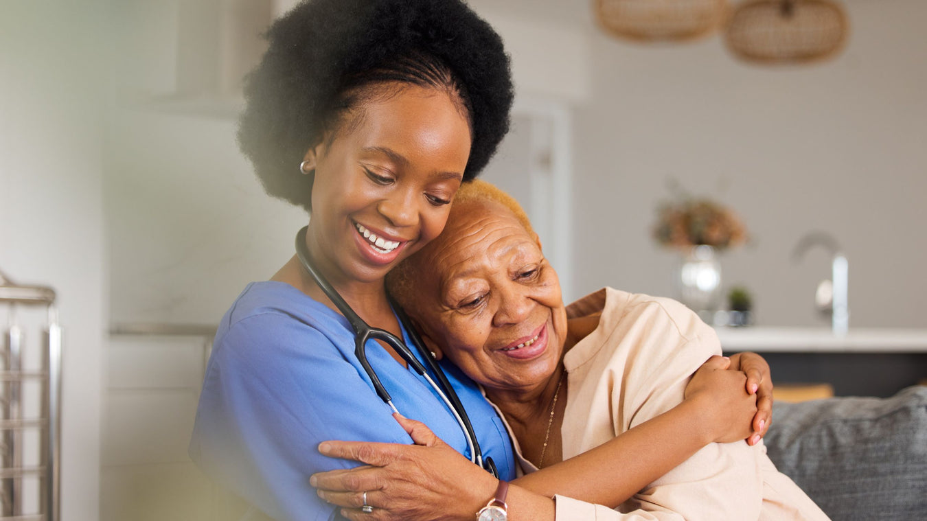 A smiling healthcare worker in blue scrubs hugs an elderly woman in a softly lit indoor setting.